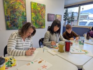 Three women sit at a white table painting flowers on watercolor paper.