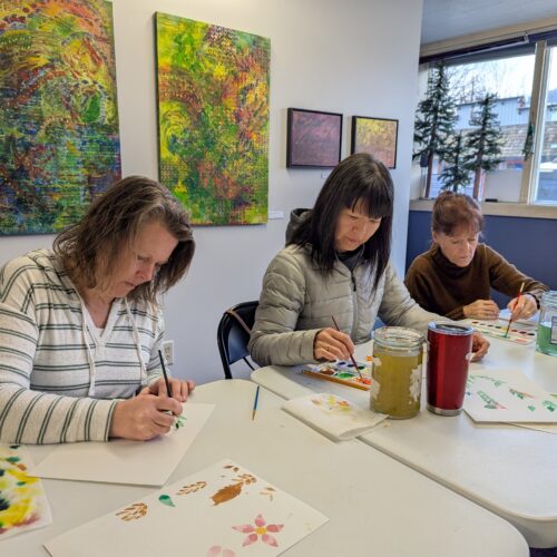 Three women sit at a white table painting flowers on watercolor paper.