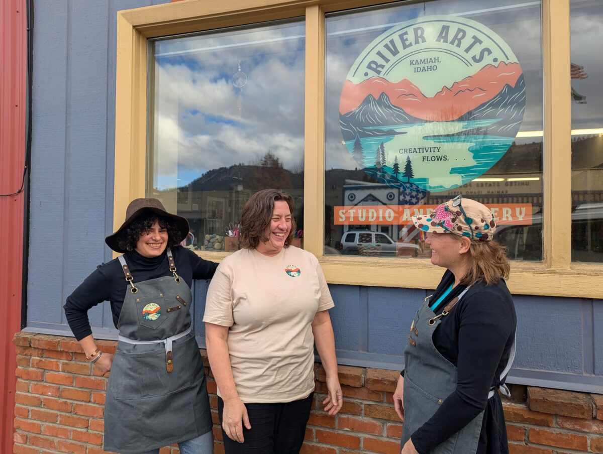 Three women stand in front of a blue and orange logo with a mountain landscape that says, "Kamiah River Arts: Creativity Flows"