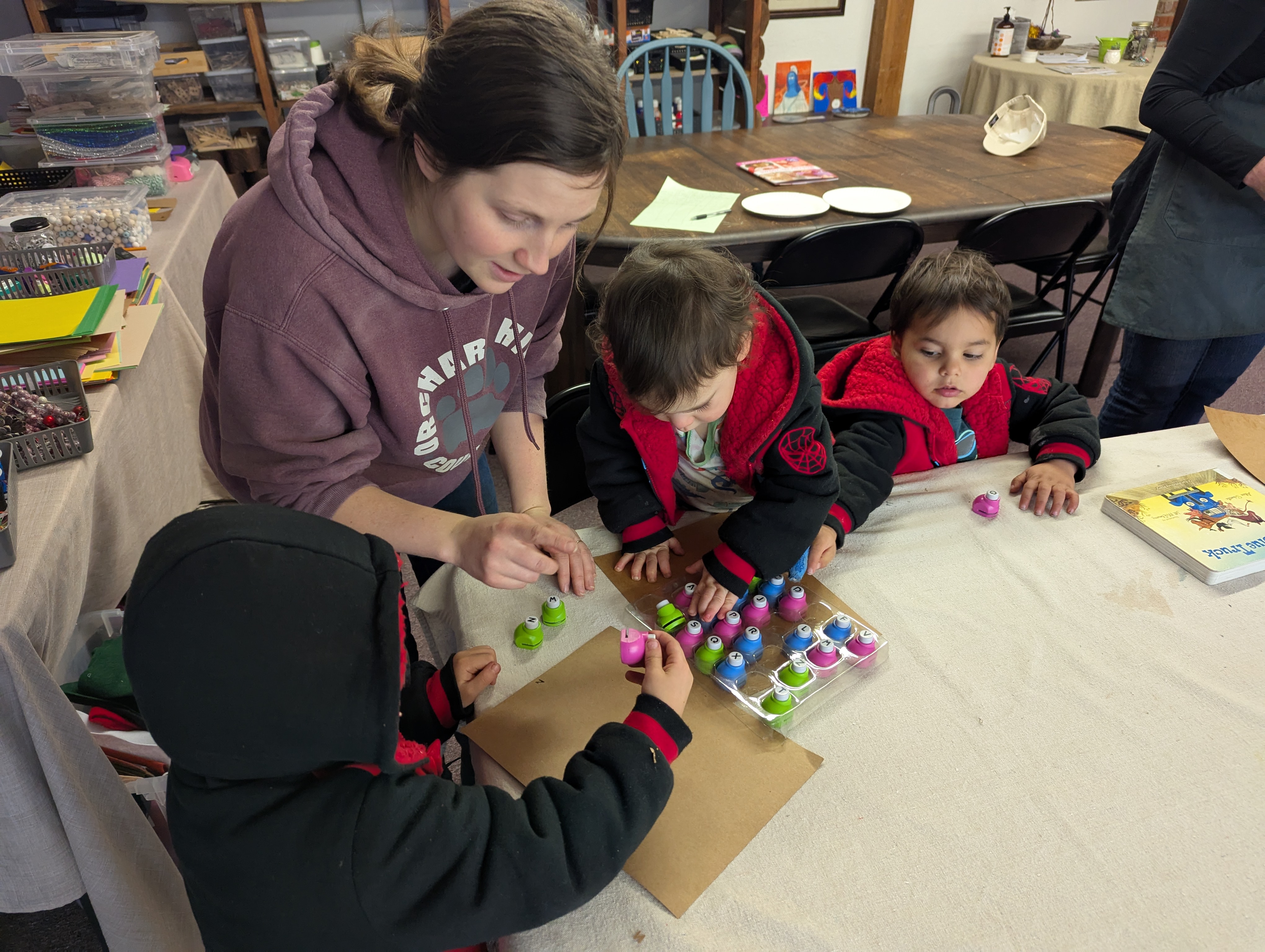 A mom in a purple sweatshirt and her three young sons sit at a table preparing to do stamping art.