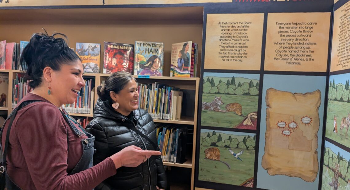 Two women with black hair look at a newly painted mural in panels with writing above each section in the children's section of the Lewiston City Library.