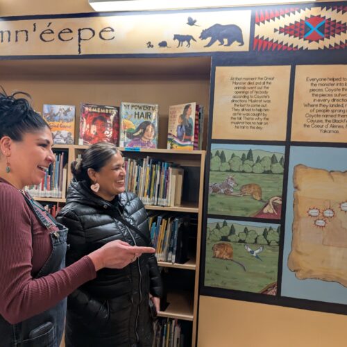 Two women with black hair look at a newly painted mural in panels with writing above each section in the children's section of the Lewiston City Library.