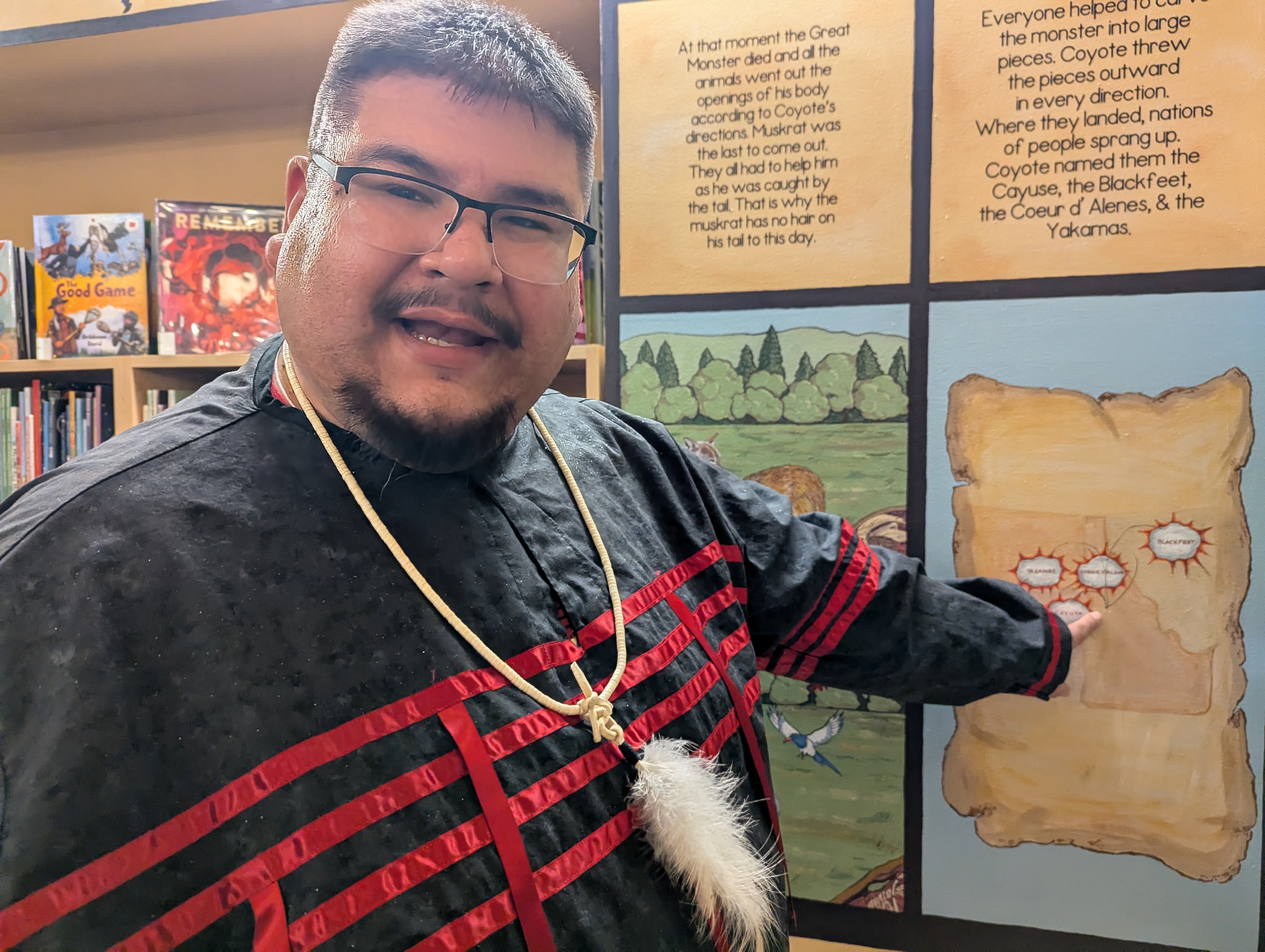 A man with black hair, goatee and glasses wearing a feather necklace points to a map on the Heart of the Monster mural. 