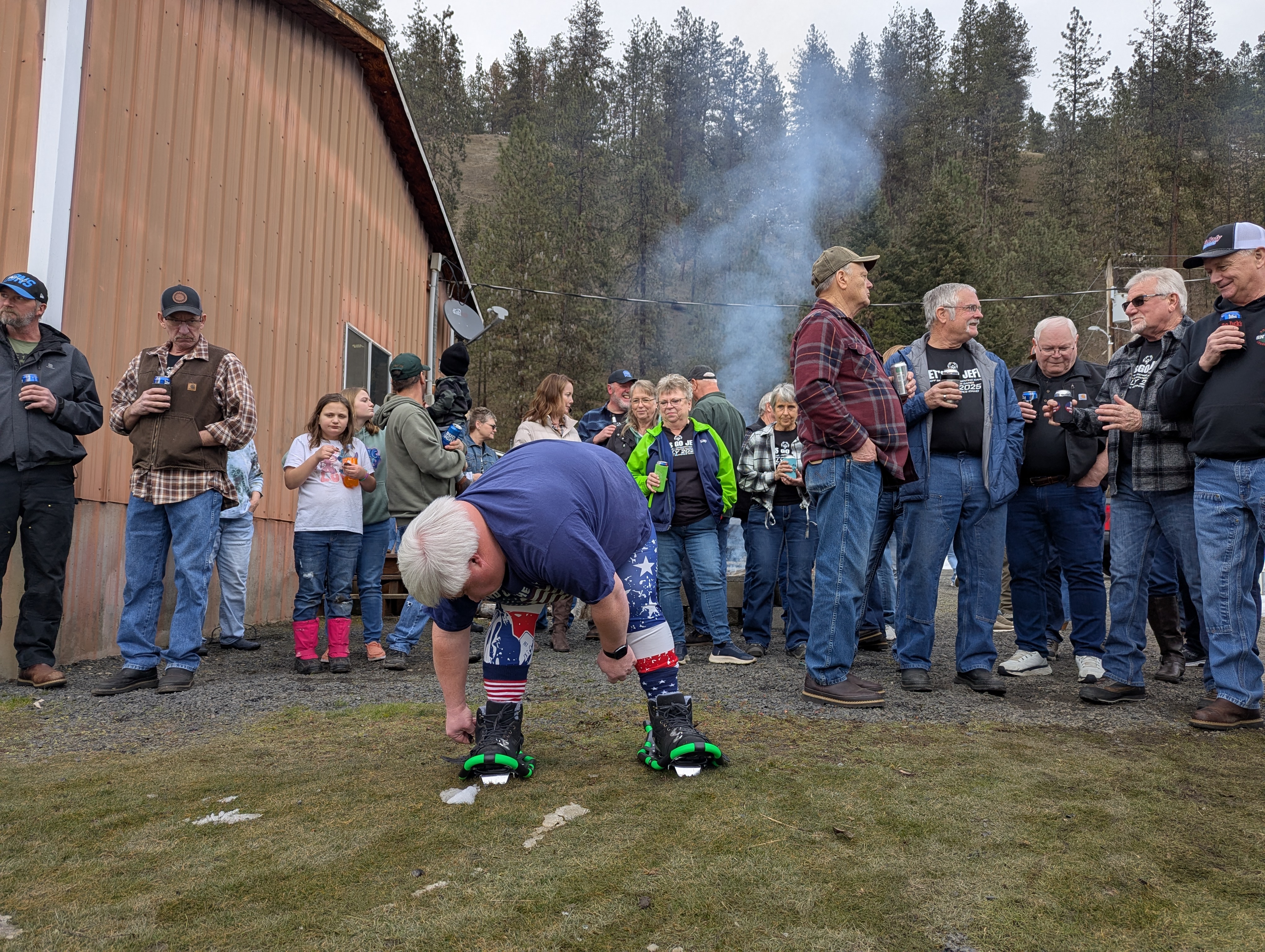 Jeff Greene straps on his black and green snowshoes on the lawn while a crowd stands behind him. 