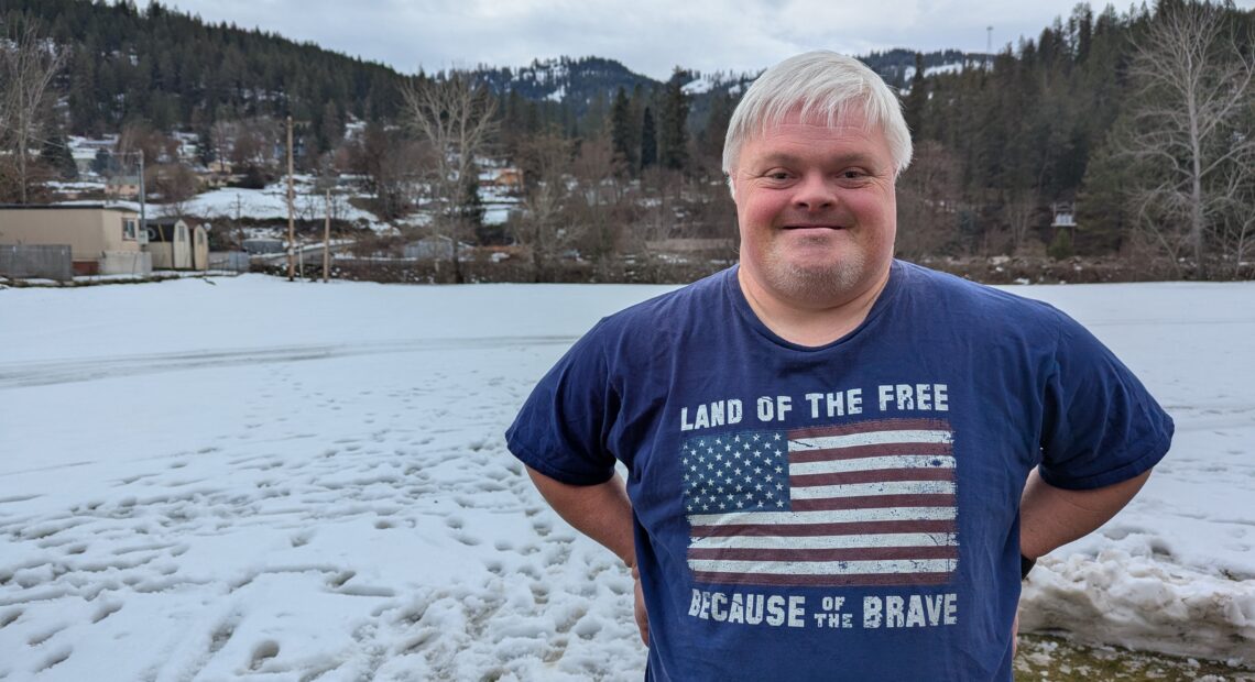 A man with Down syndrome wears a shirt with an American flag and stands in front of a snow covered yard with tree covered hills in the distance.