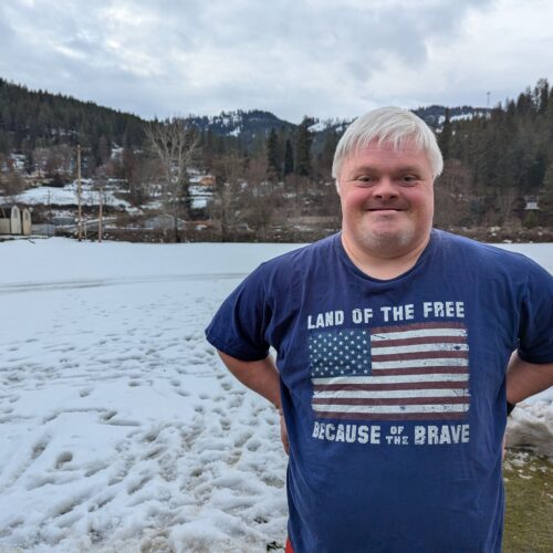 A man with Down syndrome wears a shirt with an American flag and stands in front of a snow covered yard with tree covered hills in the distance.