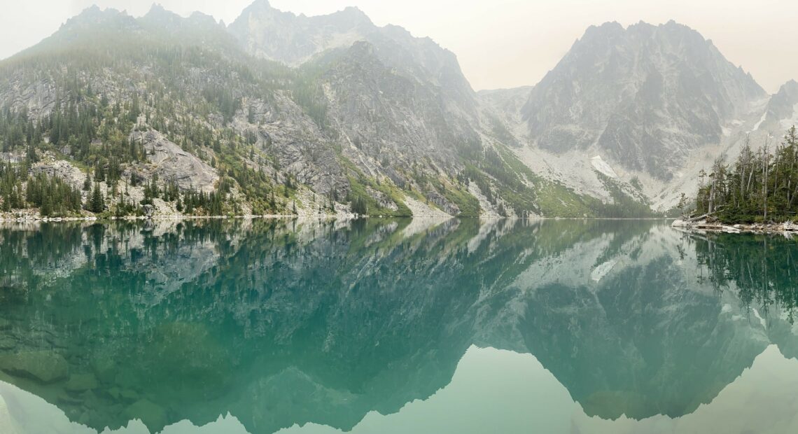 A view shows mountains dotted with trees reflected in the turquoise waters of Lake Colchuk in Washington.