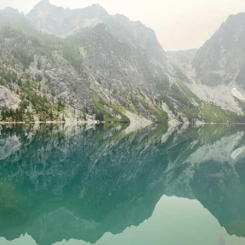 A view shows mountains dotted with trees reflected in the turquoise waters of Lake Colchuk in Washington.