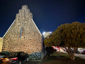 A gray stone church is next to a green round tree. The sky is dark blue. Next to the tree is a busy street. A black car is parked in front of the church