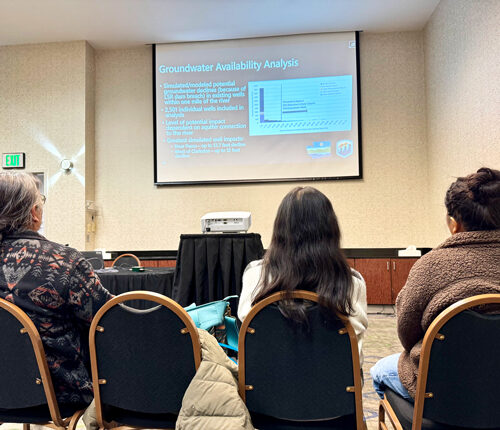 Three people sit in chairs facing the front of a room. The room has white walls. In front of the three people is a projector screen and a projector. There is a blue slide on the projector.