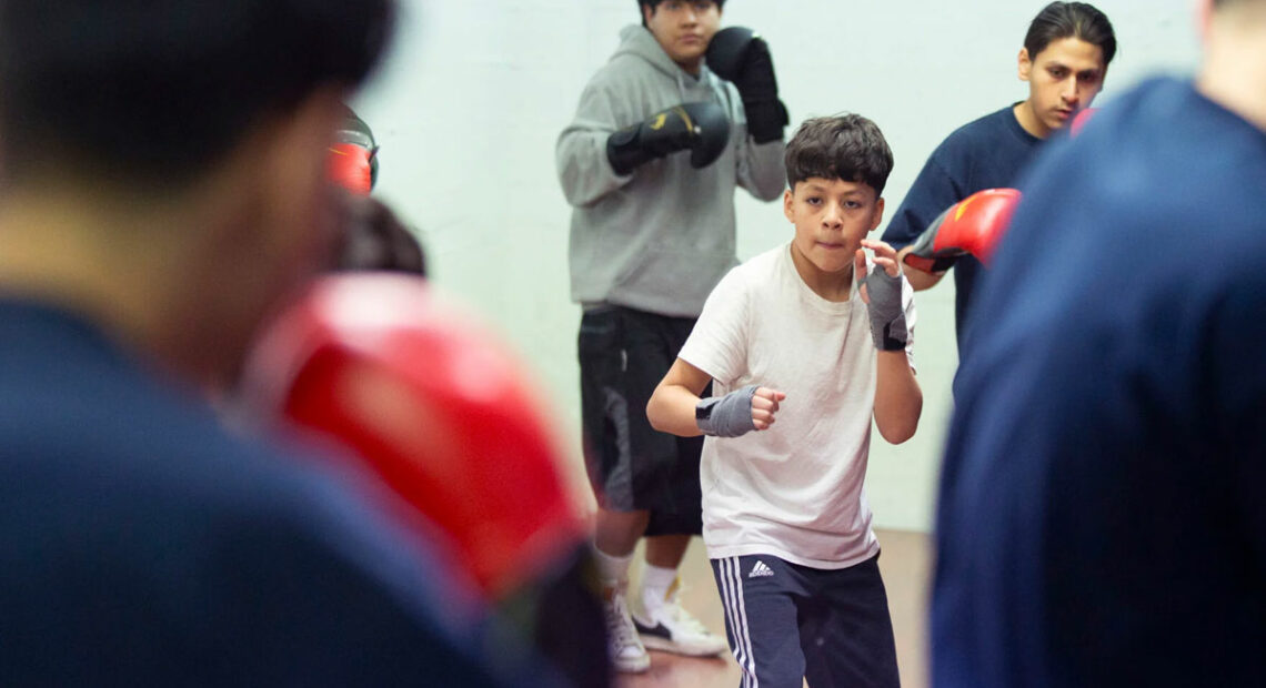 A teenager wearing a white shirt and blue pants leads a group in a boxing drill.