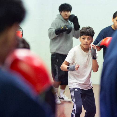 A teenager wearing a white shirt and blue pants leads a group in a boxing drill.