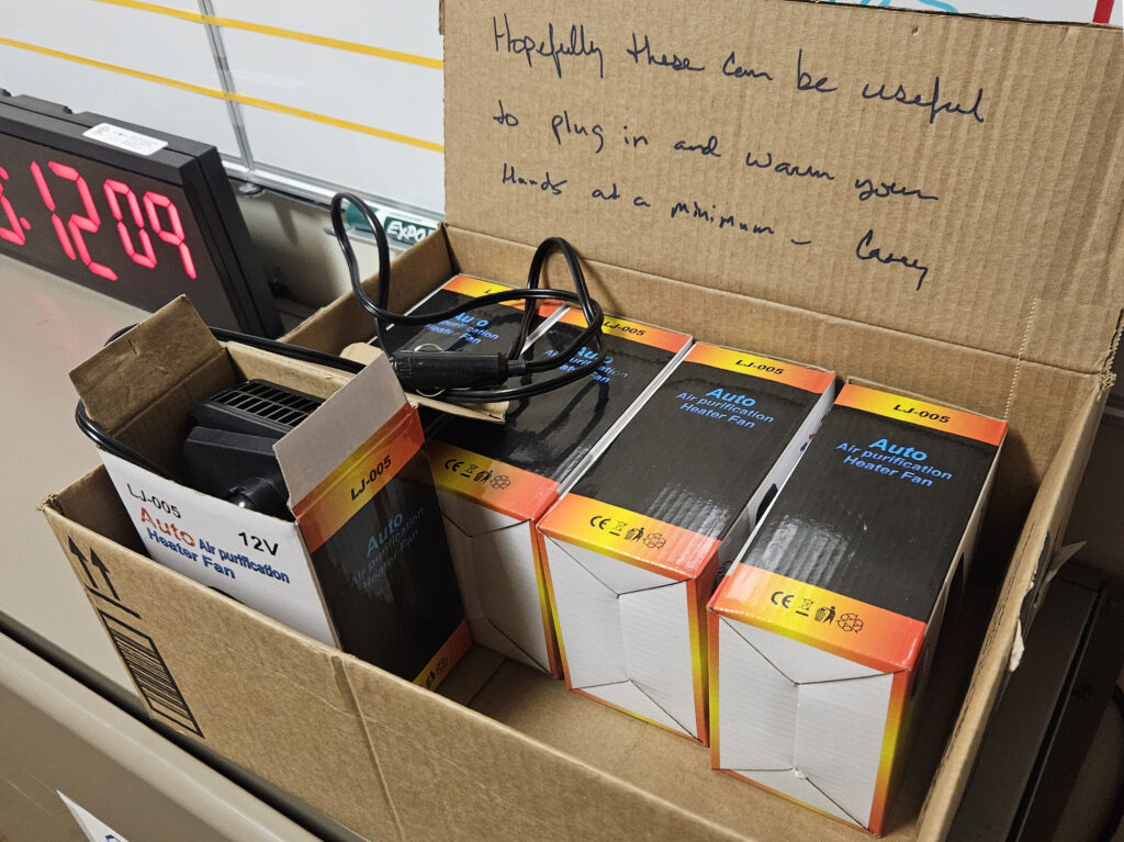 An open box of heaters sits in front of a whiteboard. A message, "Hopefully these can be useful to plug in and warm your hands at a minimum. — Casey" is written in cursive on the lid of the box.