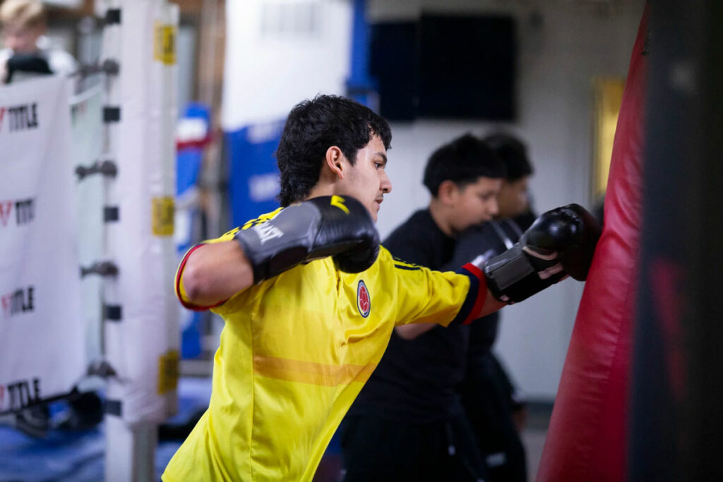 A teenager in a bright yellow shirt hits a punching bag. He is wearing black boxing gloves.