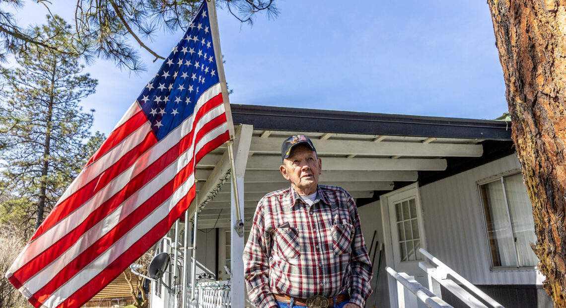 A man in a plaid shirt and glasses stands next to his white manufactured home next to a large American flag.