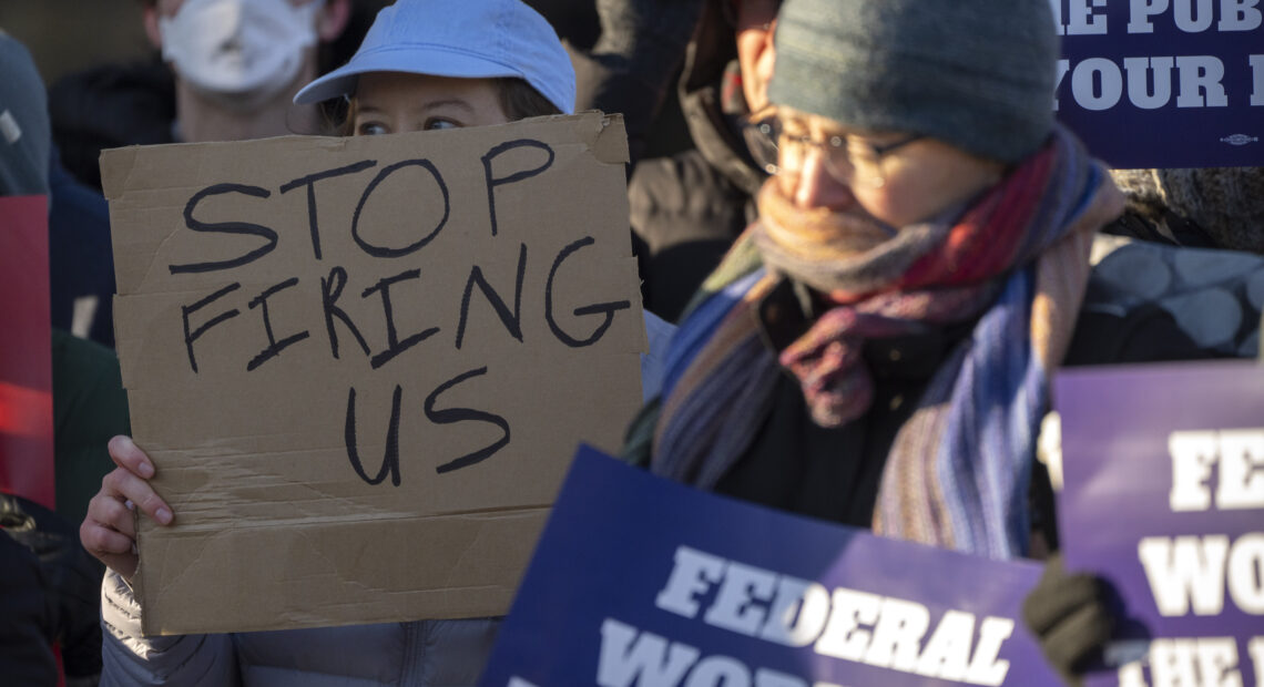 A woman in a blue hat holds a sign that reads, "stop firing us."