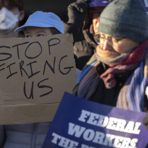 A woman in a blue hat holds a sign that reads, "stop firing us."