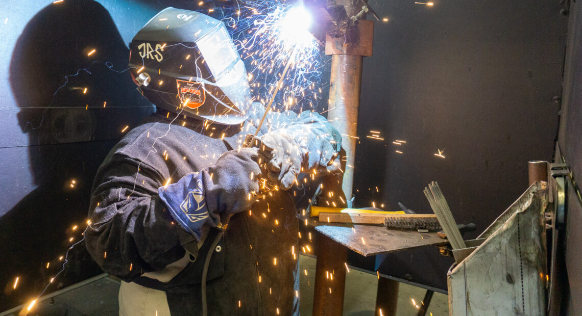 A welder in a helmet connects two pieces of metal as sparks fly.