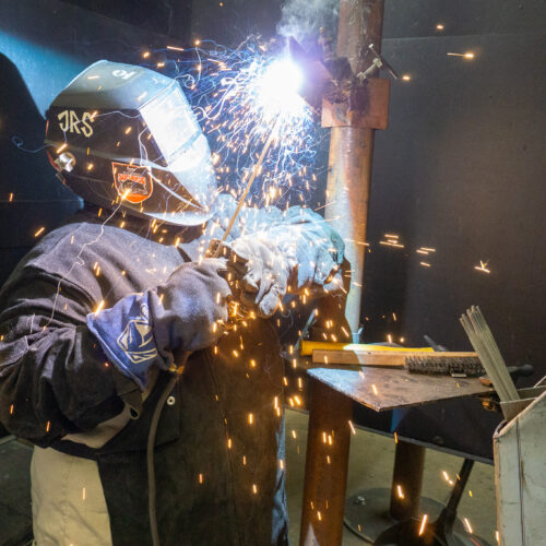 A welder in a helmet connects two pieces of metal as sparks fly.