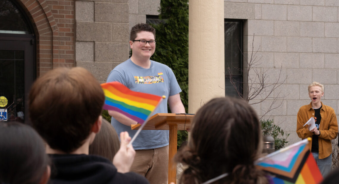 A man in a blue shirt smiles at people holding rainbow flags.