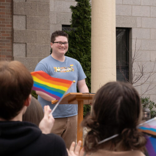 A man in a blue shirt smiles at people holding rainbow flags.