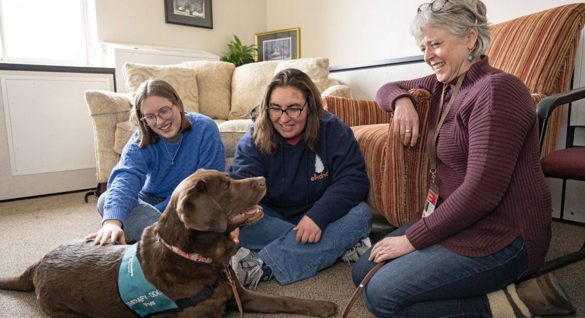 Two young students sit with a woman with gray hair against a couch. Both students are petting a chocolate lab.