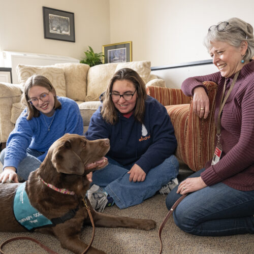 Two young students sit with a woman with gray hair against a couch. Both students are petting a chocolate lab.