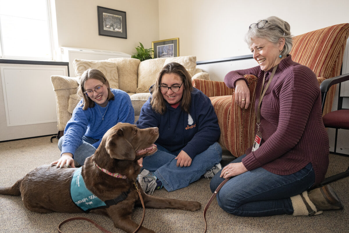 Two young students sit with a woman with gray hair against a couch. Both students are petting a chocolate lab.