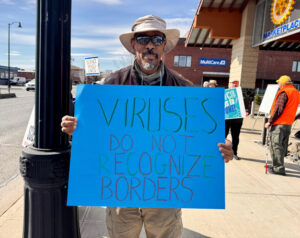 A person wearing a hat and sunglasses holds a bright blue sign that reads "viruses do not recognize borders."