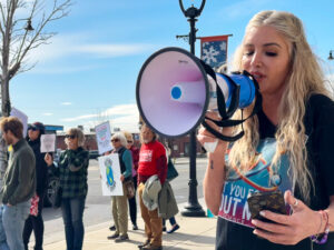 A person wearing a black shirt reads off a cell phone and speaks into a megaphone. A group of people stand in the background, holding signs.