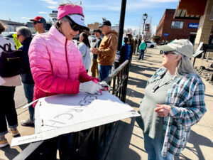 A person in a bright pink puffer jacket works on creating a sign out of an old pizza box. Another person watches their work. (Credit: Courtney Flatt / NWPB)