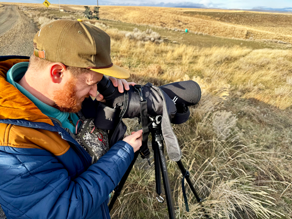 A man in an orange and blue jacket with a tan baseball hat looks through a black spotting scope that's on a black tripod. There are bunch grasses in the background on the hills.