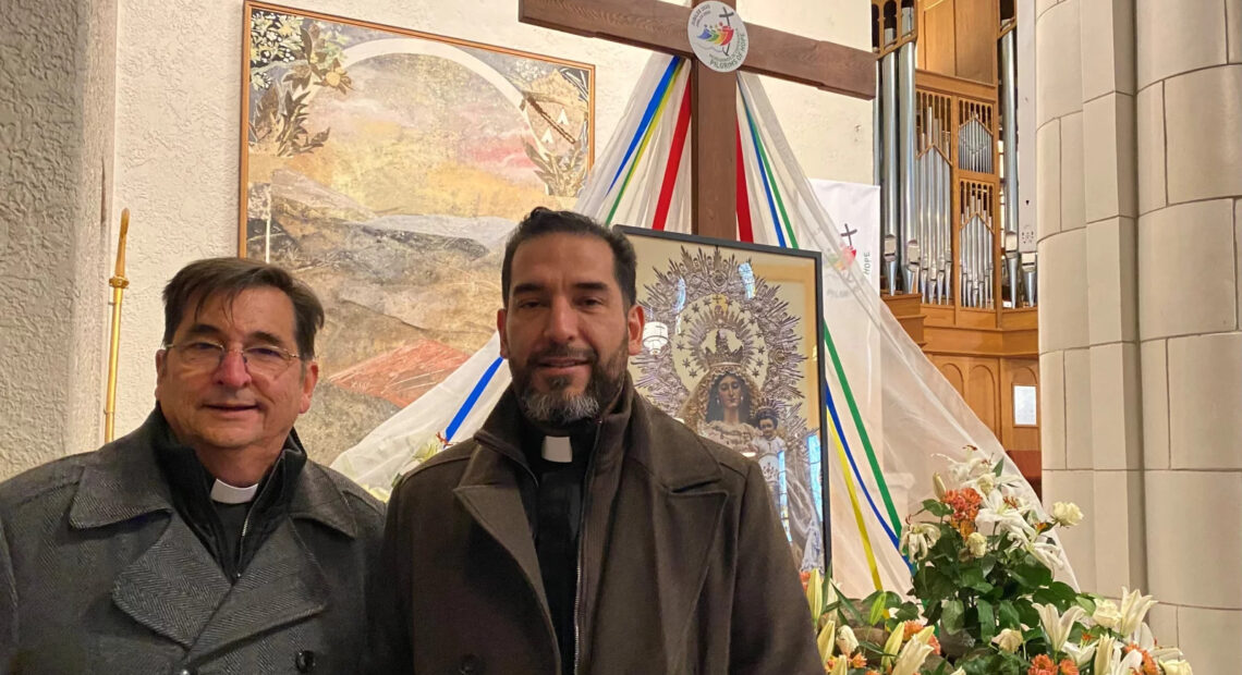 Two men stand in front of a cross. Next to the men, to the right, is a display of flowers.