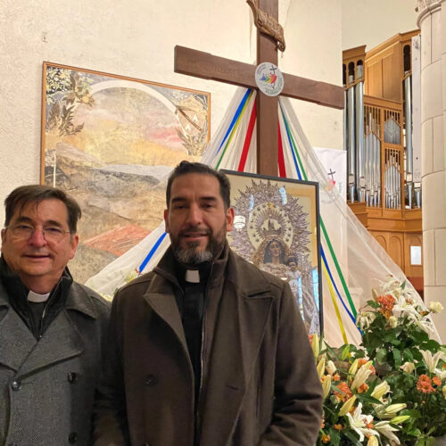 Two men stand in front of a cross. Next to the men, to the right, is a display of flowers.